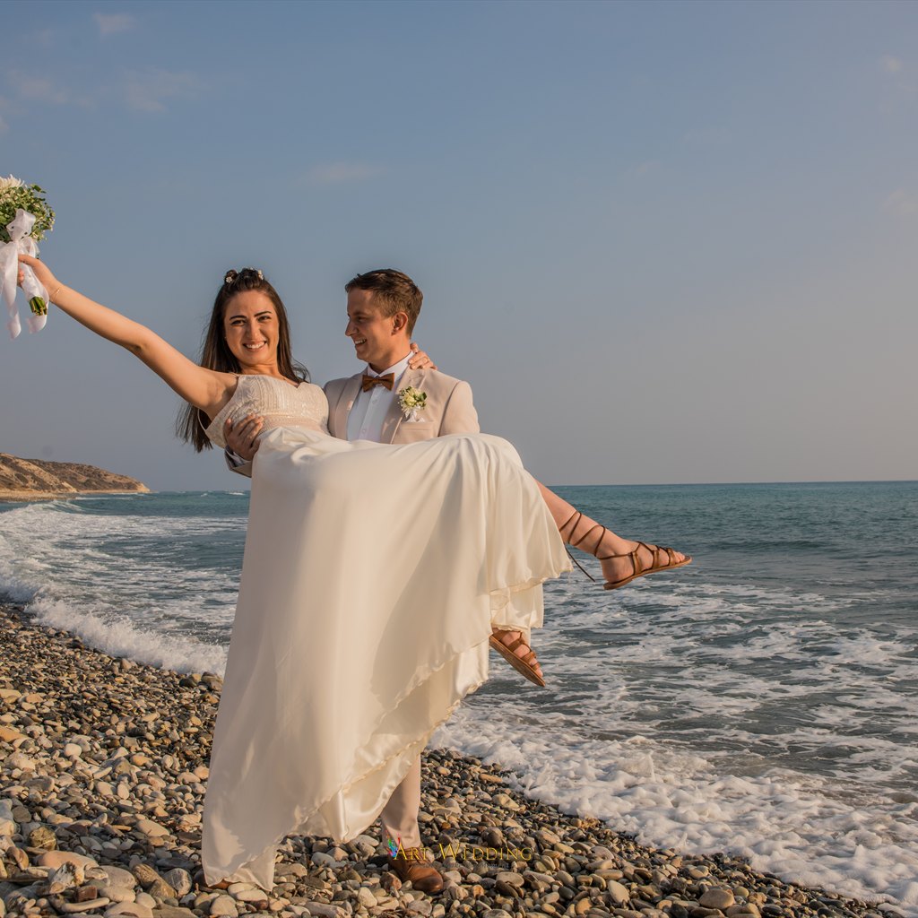 Groom carrying the bride with her bouquet raised by the sea