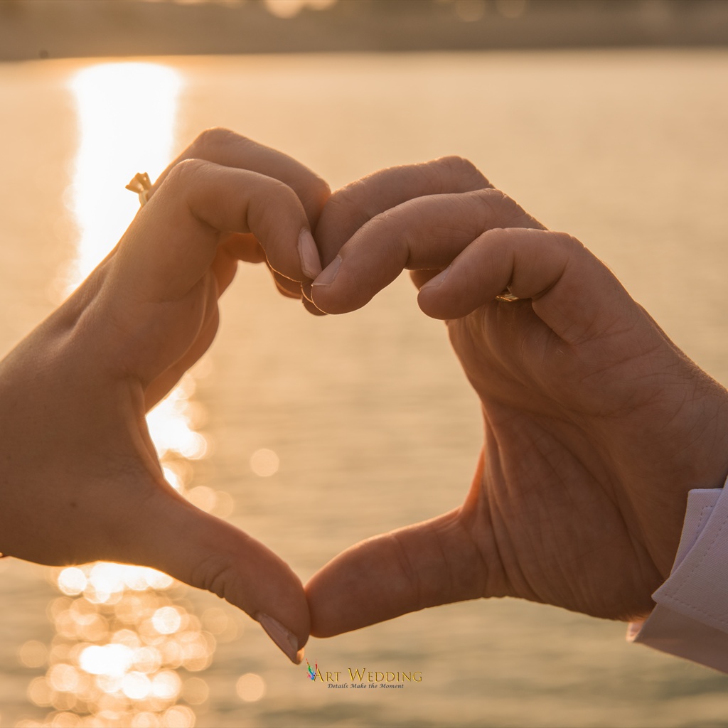 Newlyweds’ hands forming a heart against the sunset water