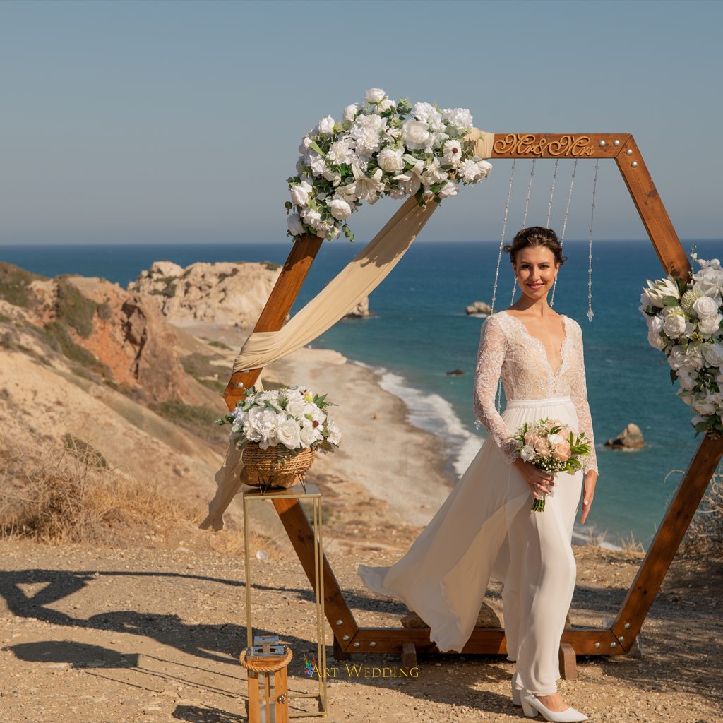 Bride holding her bouquet by an arch overlooking Petra tou Romiou