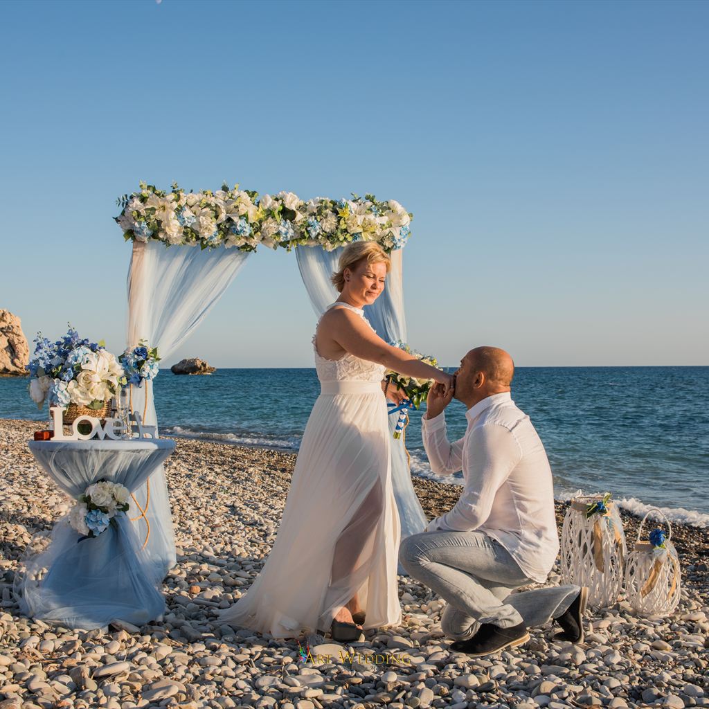 Groom kissing the bride’s hand by a blue arch on the beach