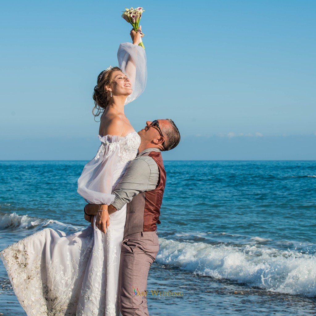 Groom lifting the bride with her bouquet against the sea waves