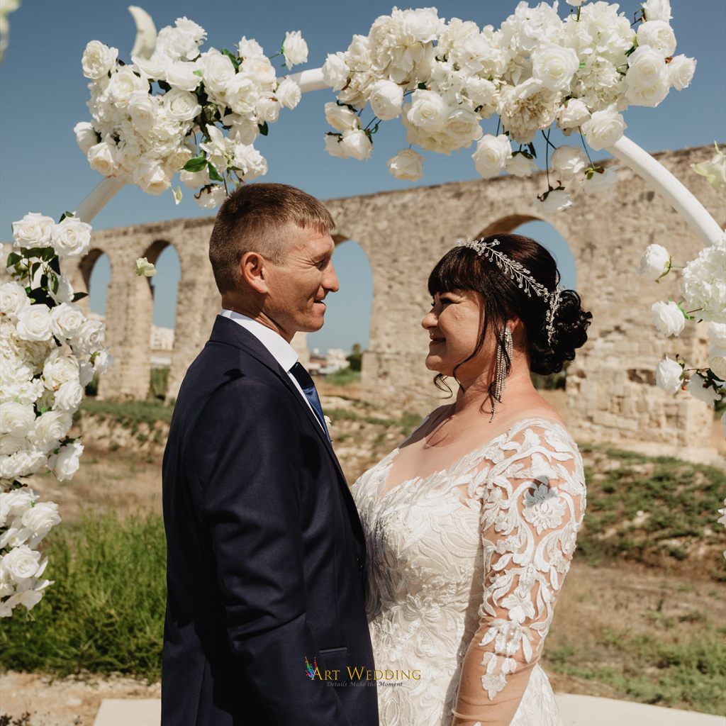 Newlyweds beneath a white floral arch with Kamares behind them