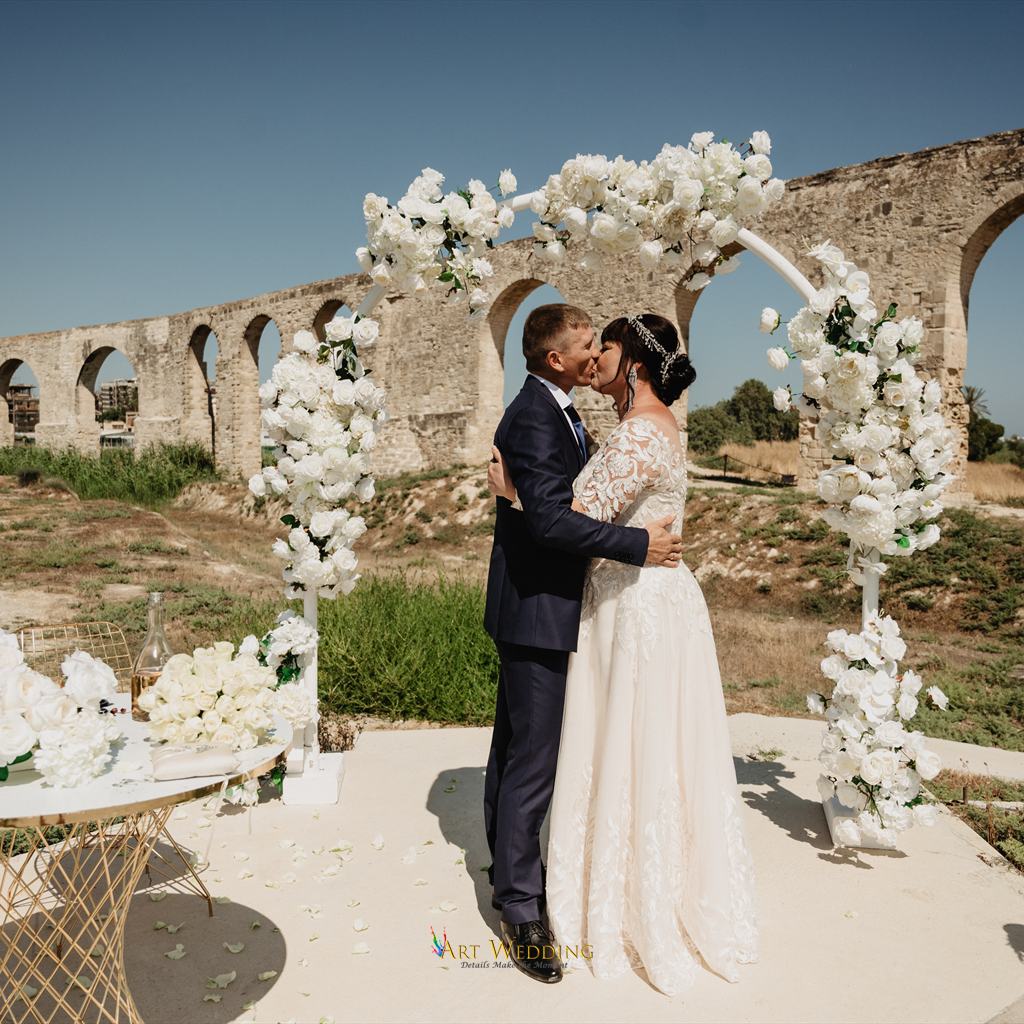 Newlyweds kissing beneath a floral arch at Kamares Aqueduct