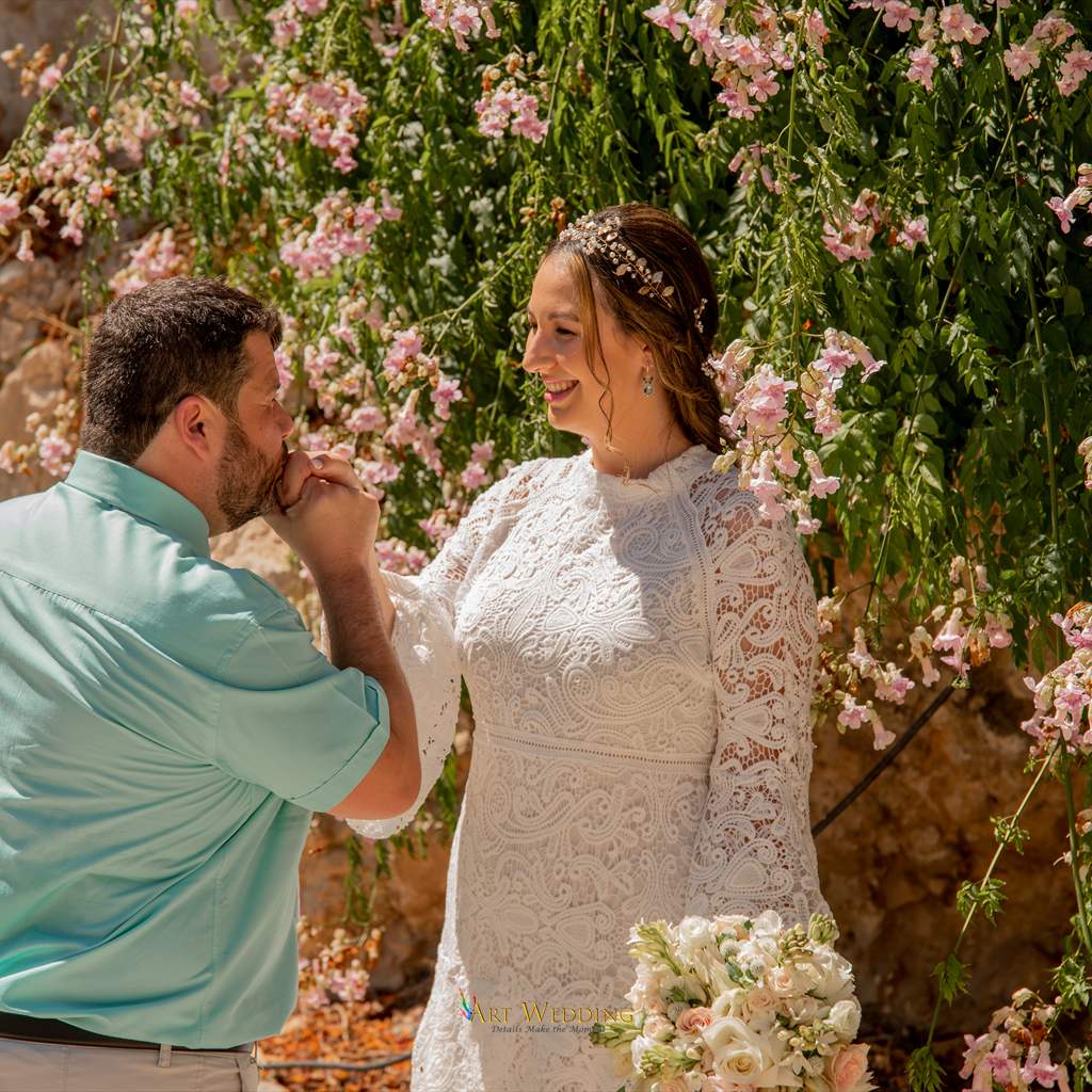Groom kissing the bride’s hand beside a flowering wall