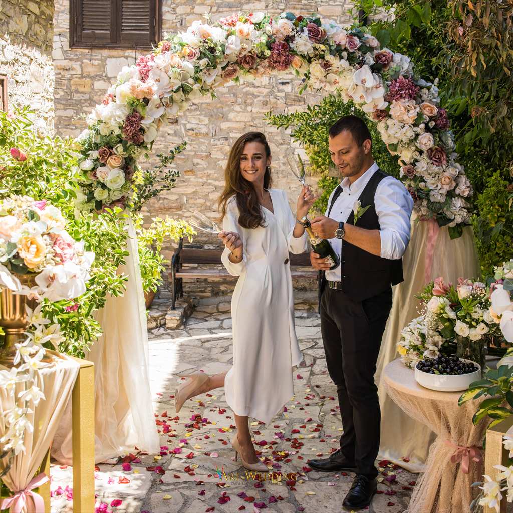 Newlyweds with champagne beneath a floral arch — Lefkara, Cyprus